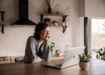 Free Young cheerful female smiling and talking via laptop while sitting at wooden table in cozy kitchen Stock Photo