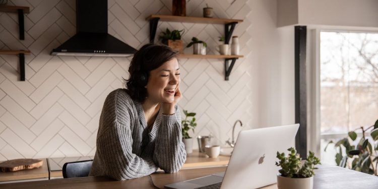 Free Young cheerful female smiling and talking via laptop while sitting at wooden table in cozy kitchen Stock Photo
